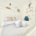Habitación luminosa con cama doble, toallas blancas enrolladas y decoración minimalista en tonos claros.