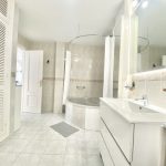 Baño moderno con bañera redonda, lavabo blanco y espejos, decorado con azulejos claros y armarios de almacenamiento.
