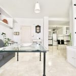 Sala de estar moderna con mesa de cristal y sofá gris, decorada con plantas y estanterías blancas, ideal para un ambiente acogedor.