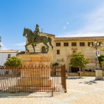 Estatua de un caballero montado en un caballo en una plaza, rodeada de edificios históricos y palmeras.