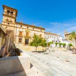 Vista de una plaza histórica en Andalucía con edificios de estilo andaluz y vegetación, ideal para turismo cultural y arquitectura.